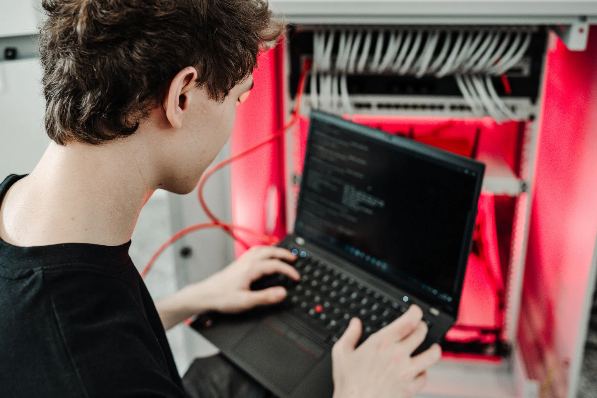 IT technician working on a laptop in front of an open server rack with red cabling